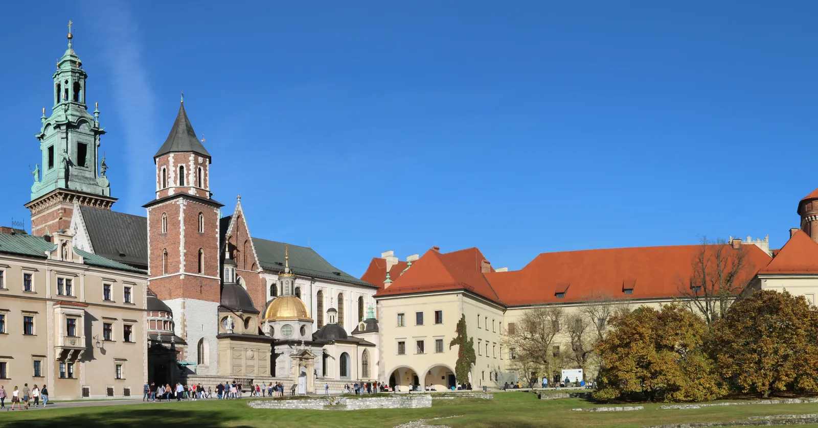 Wawel Cathedral and the castle wing beside it on Wawel Hill in Kraków, a green-capped clock tower and a red brick belfry above a gold-domed chapel, with the castle's long orange-tiled roof running off to the right