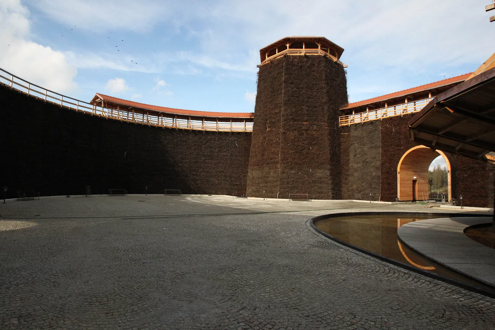Inside the brine graduation tower at Wieliczka, a tall curved wall packed with dark blackthorn brushwood curving around a paved yard, with an octagonal timber viewing tower rising at its centre