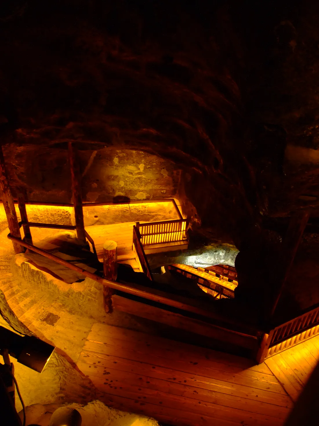 A timber staircase descending through a chamber in the Wieliczka salt mine, heavy log railings and wooden treads lit warm yellow against dark rock walls