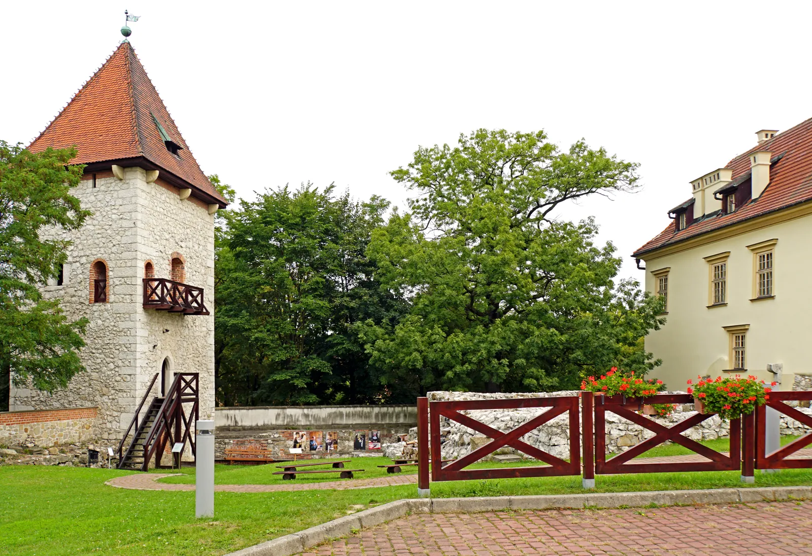 The Saltworks Castle in Wieliczka, a square stone tower with a red tiled roof and an external timber gallery, the pale castle wing beside it and excavated foundations in the lawn between them