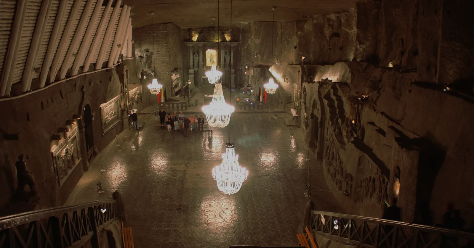 St Kinga's Chapel deep in the Wieliczka salt mine, a large rock-cut hall with three crystal chandeliers hanging over a polished floor, carved reliefs along the walls and a lit altar at the far end
