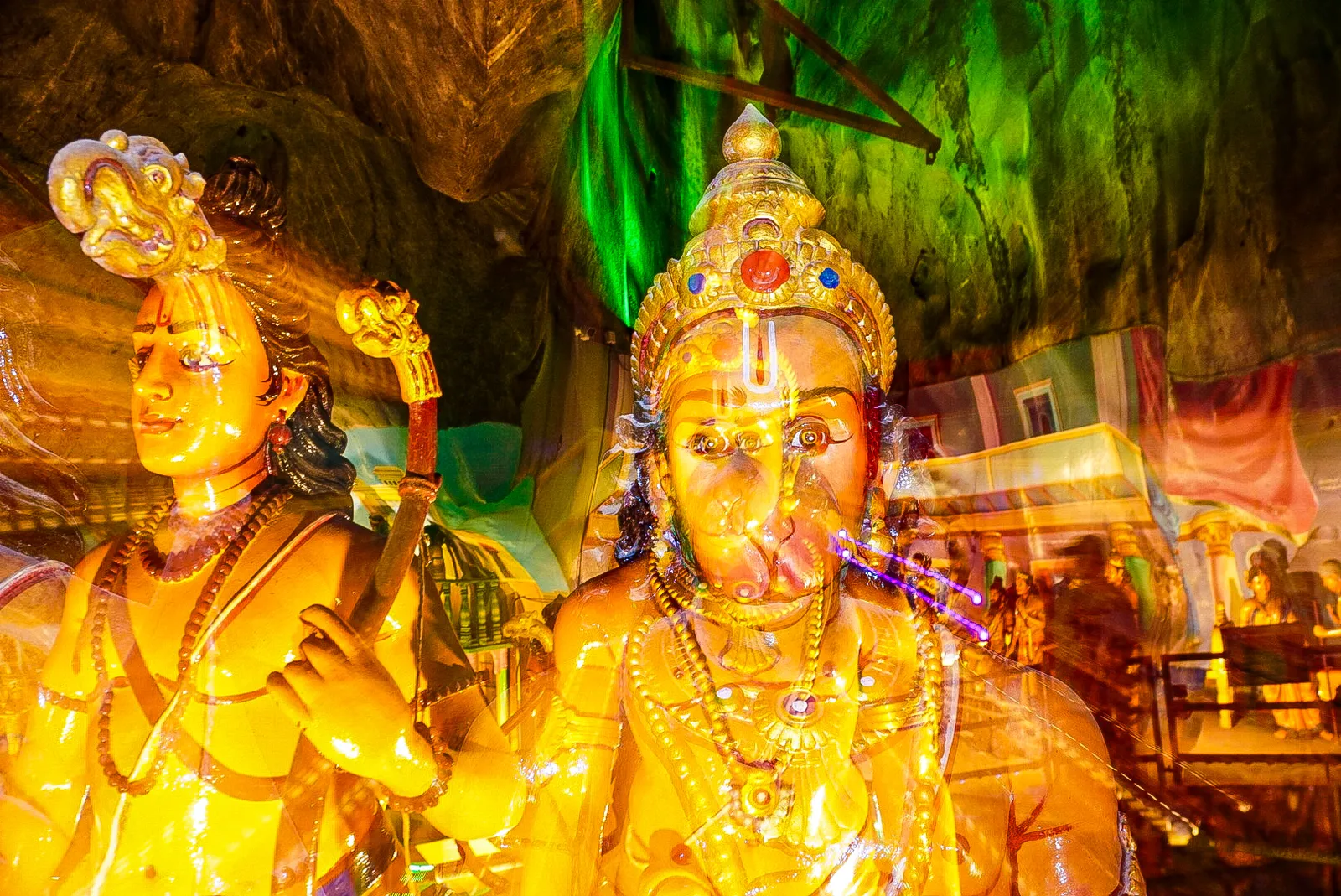 Painted statues of Hanuman and Rama in the Ramayana Cave at Batu Caves, gilded figures lit green against the rough cave wall behind them
