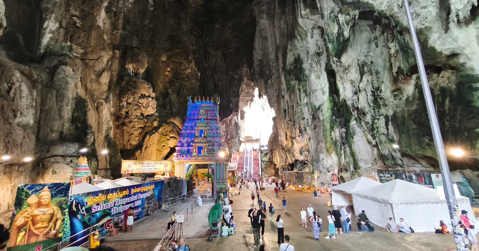 The main chamber of the Temple Cave at Batu Caves, a painted Hindu shrine tower standing on the cave floor beneath ribbed limestone walls hundreds of feet high, with visitors crossing the open ground in front