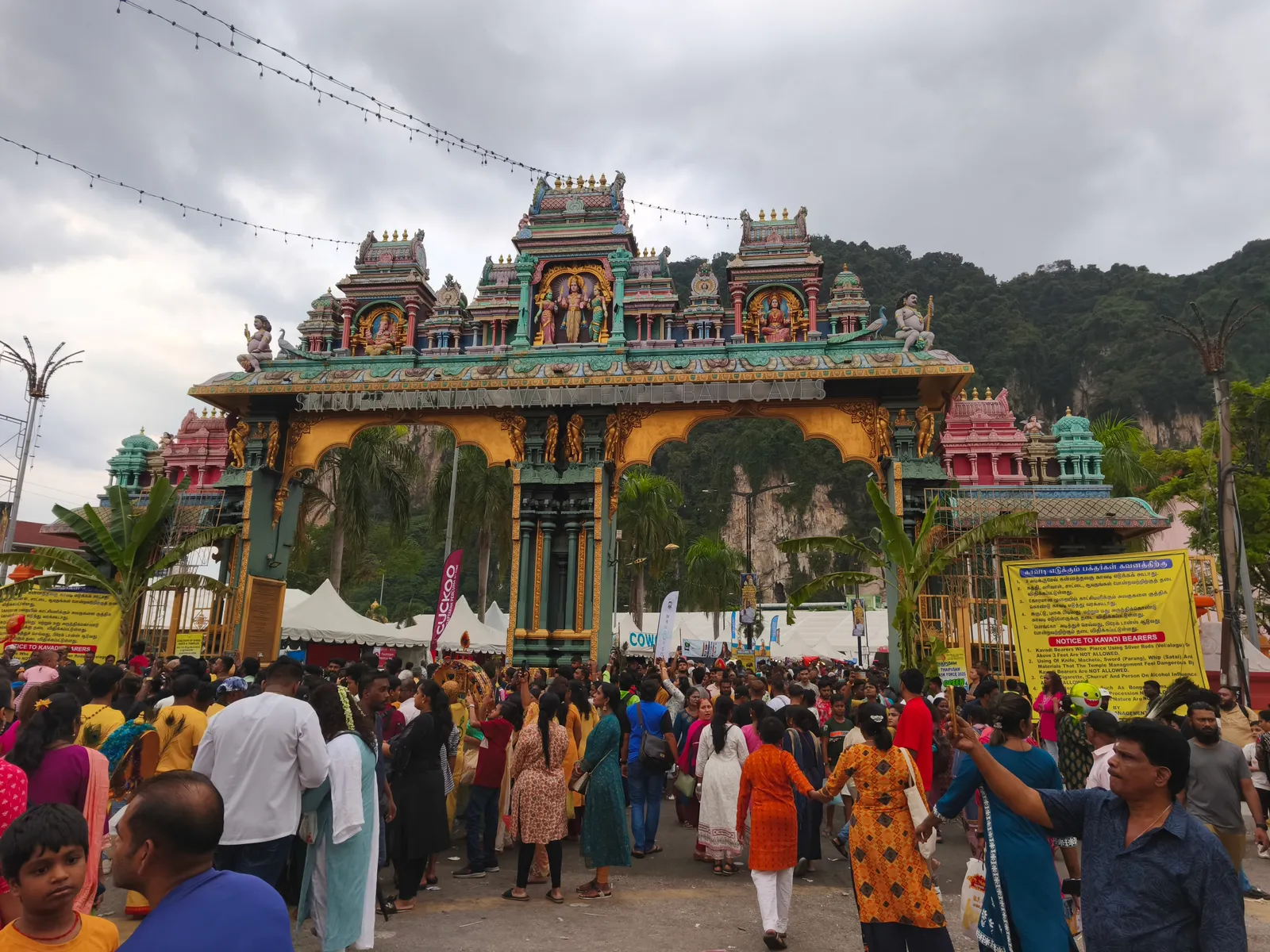 The painted gateway tower of Sri Subramaniar Swamy Temple at the foot of Batu Caves during Thaipusam, dense crowds filling the forecourt with the limestone hill rising behind