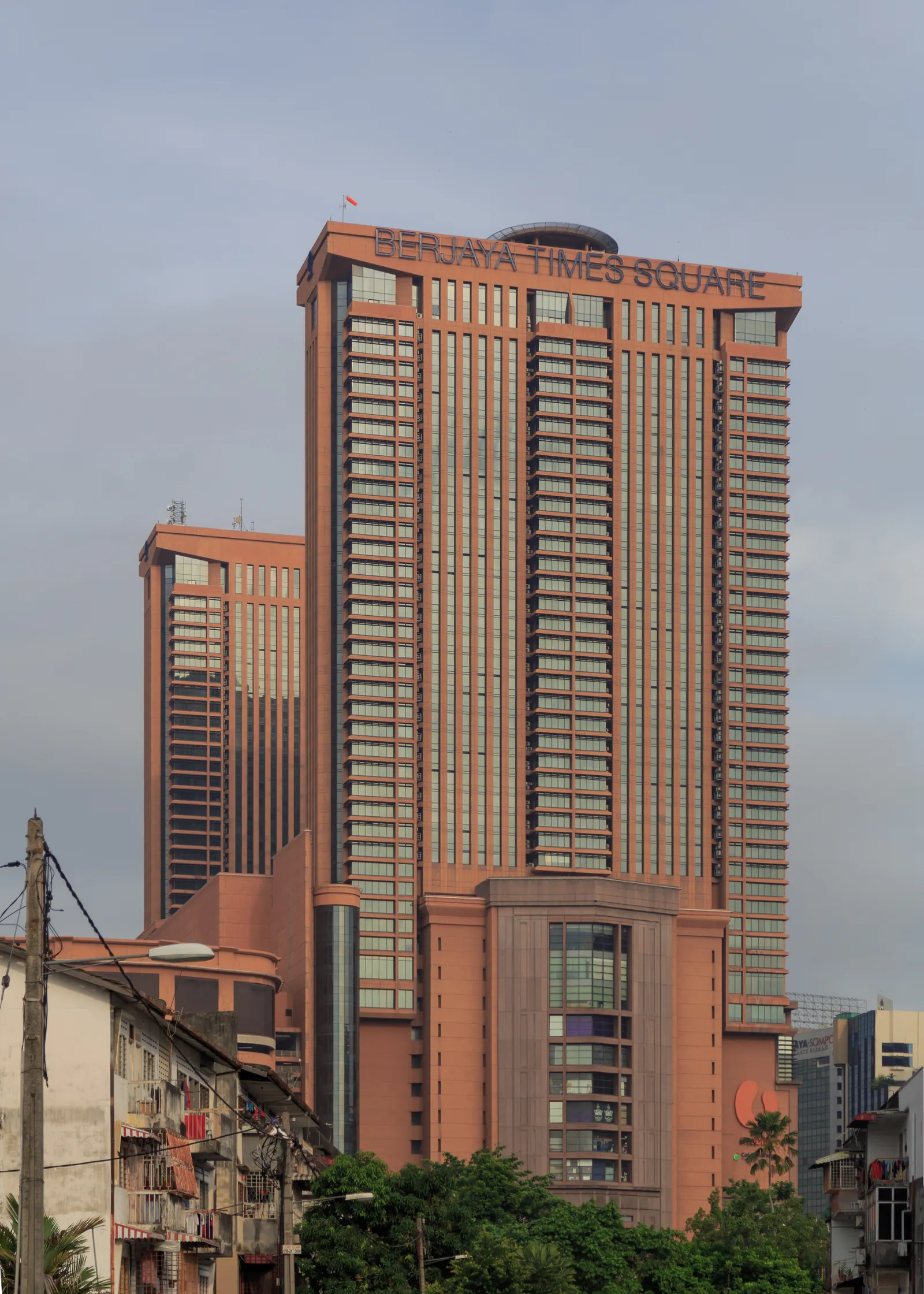 The twin red-brown towers of Berjaya Times Square in Kuala Lumpur rising above low shophouses, the building's name lettered across the roofline