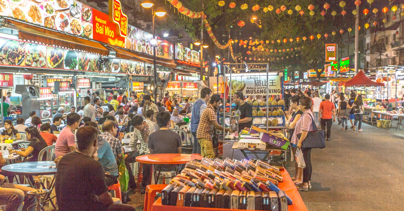 Jalan Alor in Kuala Lumpur after dark, paper lanterns strung above the road, plastic tables filling the carriageway and lit stall fronts selling fruit and grilled food down both sides