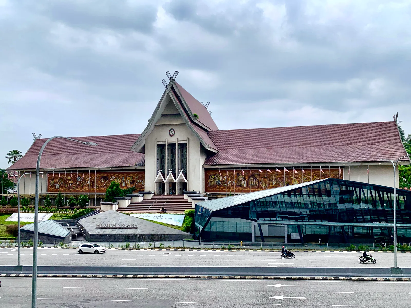 The National Museum of Malaysia in Kuala Lumpur behind its forecourt, a long low building under a steep Minangkabau roof with mosaic murals running along the upper wall on either side of the entrance
