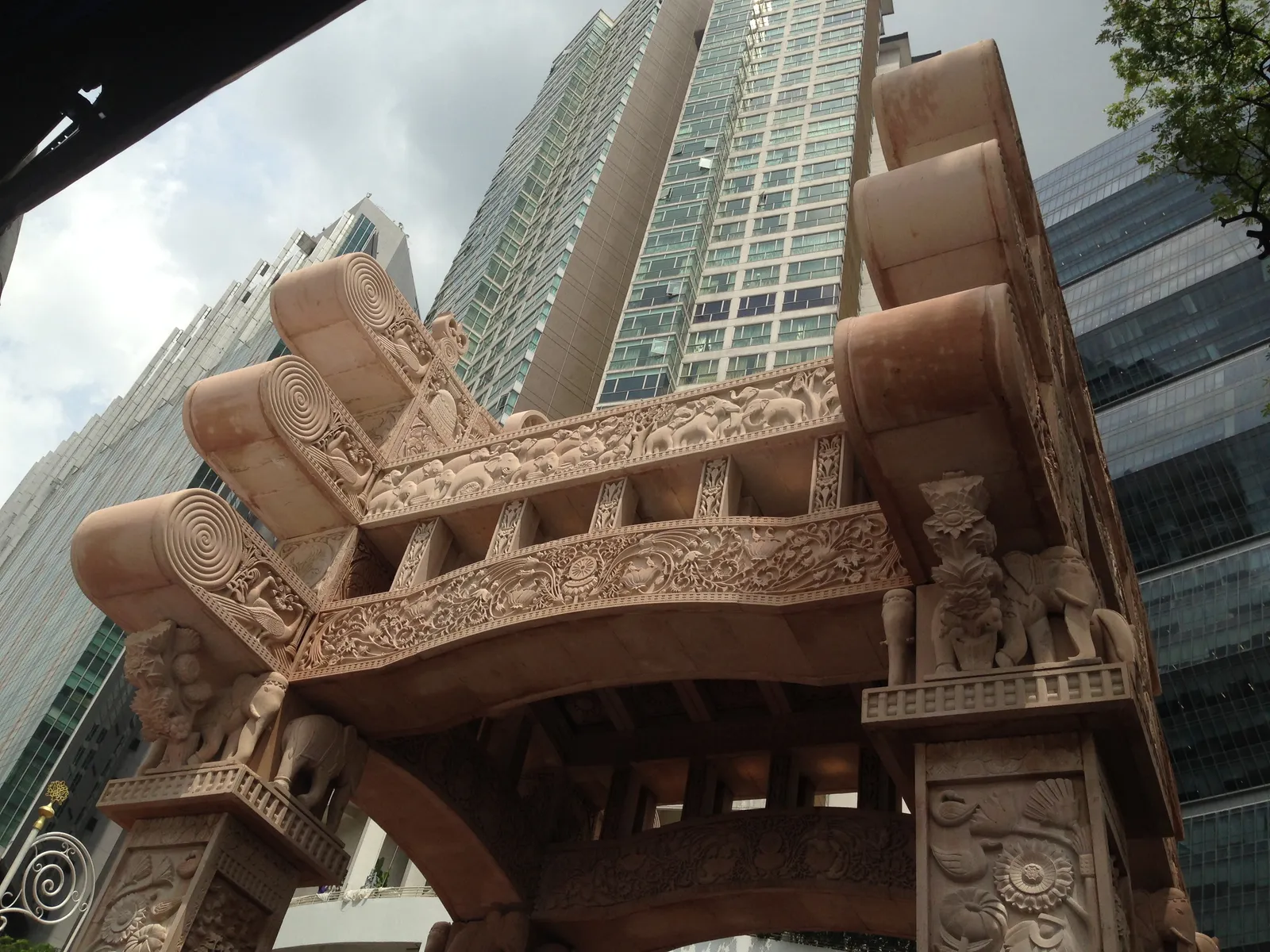 The carved sandstone Torana Gate in Brickfields, Kuala Lumpur, its three curved tiers of relief panels and elephant figures seen from below against surrounding high-rise towers