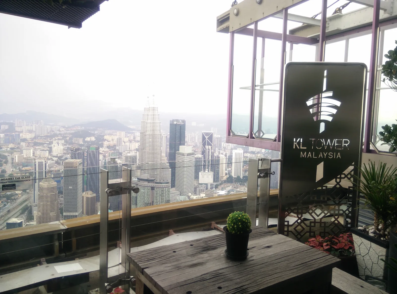 The open-air Sky Deck at Menara Kuala Lumpur, a low table and railing at the edge with the Petronas Twin Towers and the rest of the city spread out far below through the haze