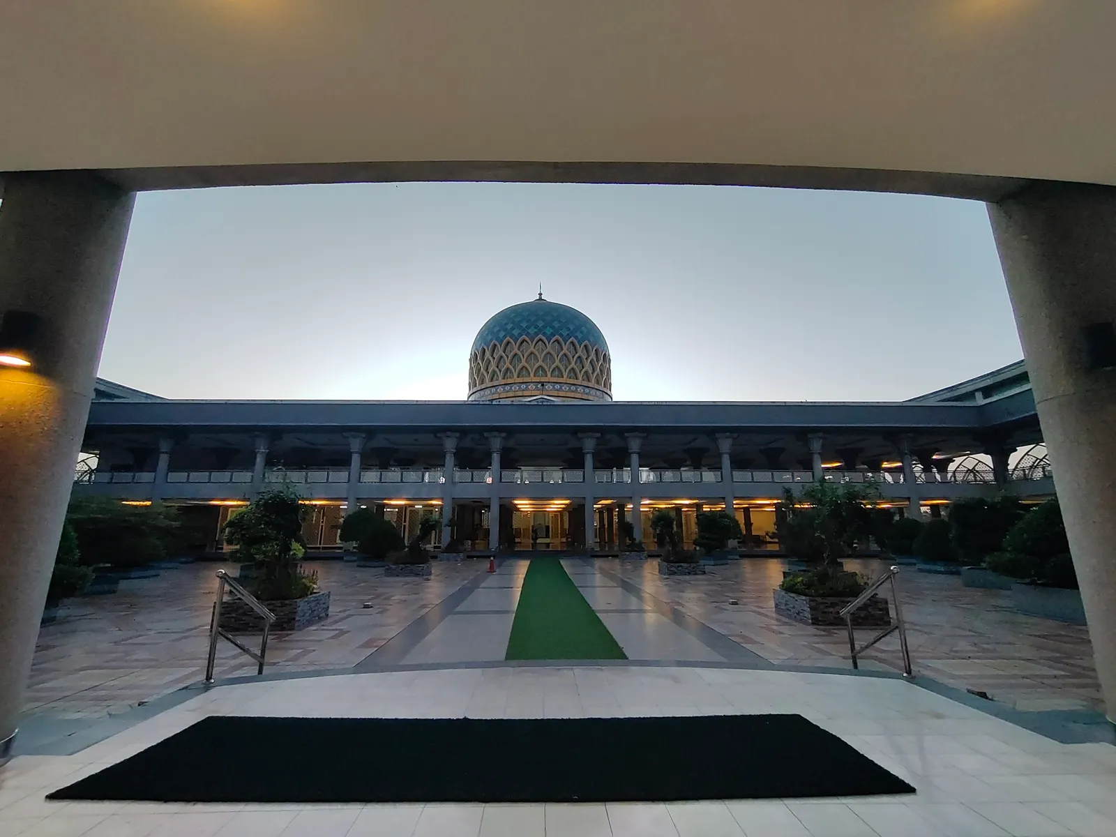 The blue and gold tiled dome of Sultan Abdul Samad Mosque at Sepang seen across its paved courtyard, framed by the arch of the entrance colonnade at dusk