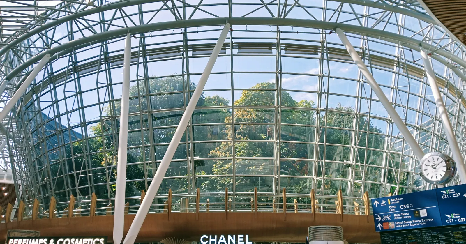 The glazed dome inside the satellite terminal at Kuala Lumpur International Airport, a stand of rainforest trees growing behind the glass above a row of duty-free shopfronts