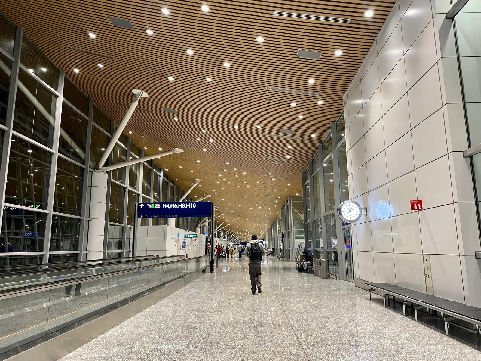 A long concourse in Terminal 1 at Kuala Lumpur International Airport under a slatted timber ceiling, glass walls on one side and a few passengers walking the length of it