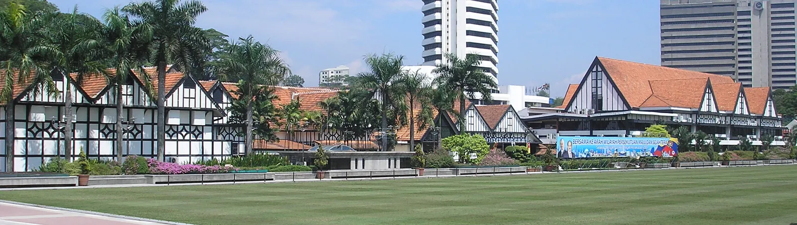 The long black-and-white mock-Tudor frontage of the Royal Selangor Club seen across the lawn of Merdeka Square in Kuala Lumpur, palms along the edge of the grass