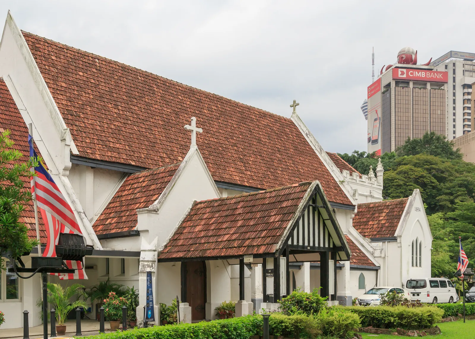 St Mary's Cathedral in Kuala Lumpur, a white gabled church under a deep red tiled roof with a covered porch, an office tower rising behind the trees