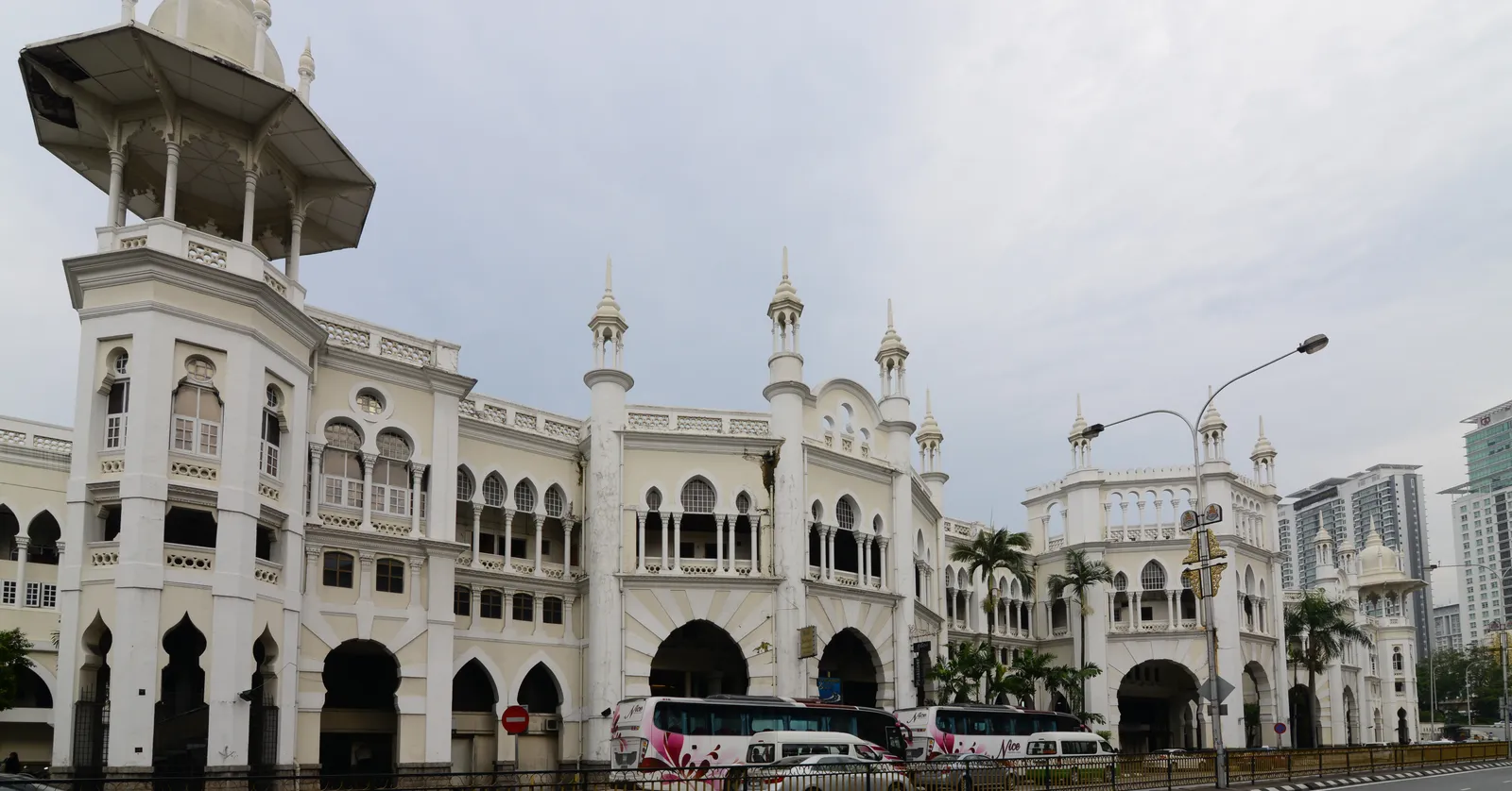 The white frontage of Kuala Lumpur railway station, a long arcade of horseshoe arches under small domed pavilions and slender minarets, with modern towers behind it
