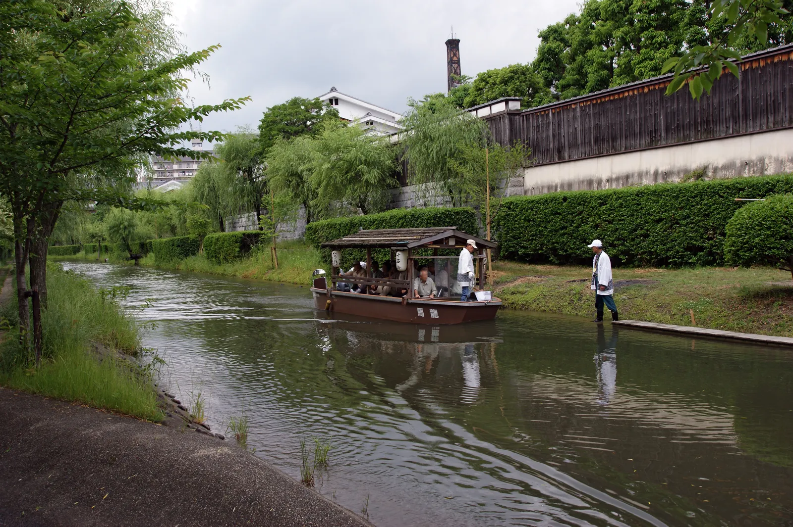 A flat-roofed wooden boat carrying passengers along a narrow willow-lined canal in Fushimi, Kyoto, with the long white and timber wall of a sake warehouse running along the far bank