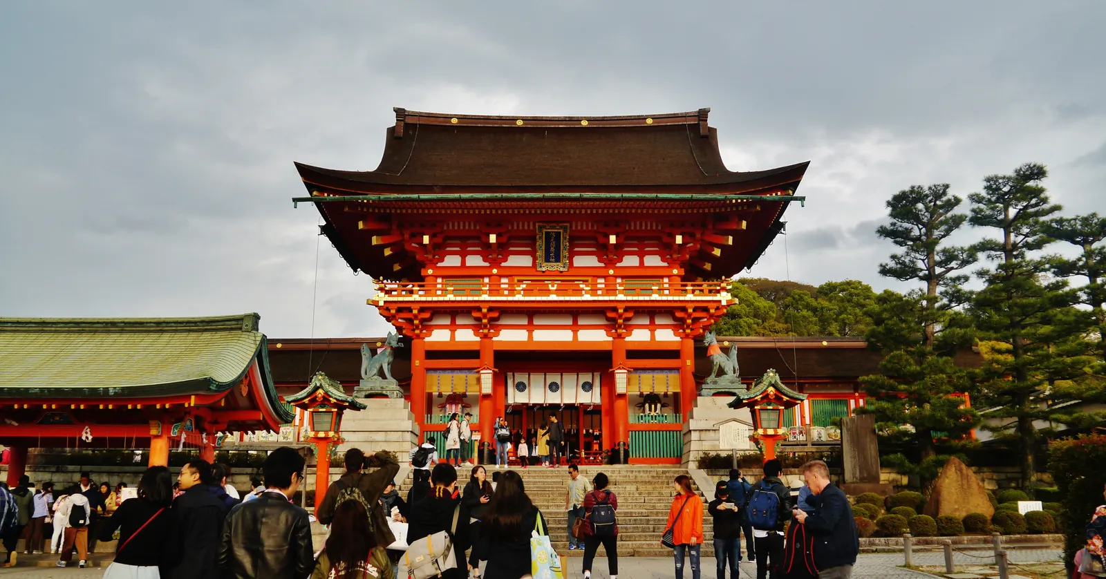 The two-storey vermilion Romon gate at Fushimi Inari shrine in Kyoto, its bright red timbers and dark tiled roofs rising above a flight of steps, with lower shrine buildings and pines on either side
