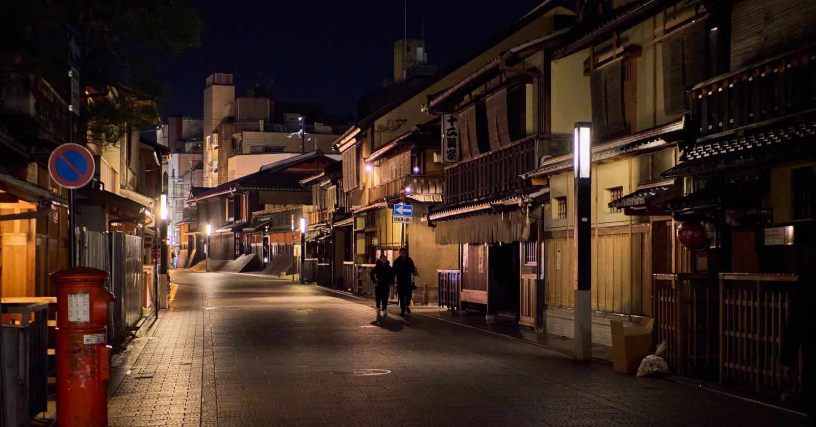 A street in Kyoto's Gion district after dark, two-storey wooden buildings with slatted timber screens and lit lanterns down both sides, and two figures walking along the empty paving between them