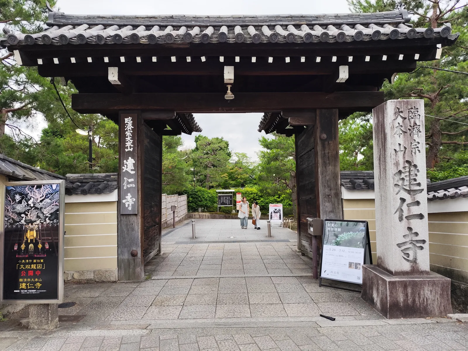 The wooden entrance gate to Kenninji temple in Kyoto, a tiled roof over dark timber posts between plastered walls, with a tall inscribed stone pillar beside it and a path leading into the grounds