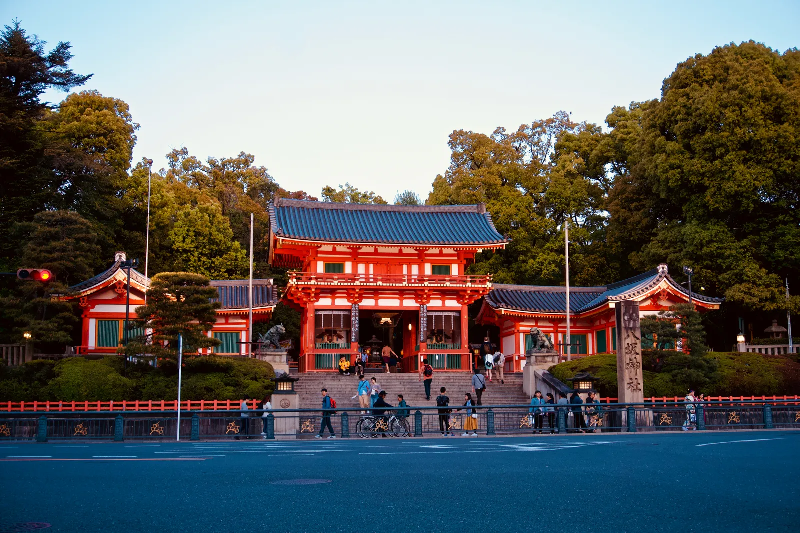 The vermilion two-storey gate of Yasaka Shrine in Kyoto seen from the street below in evening light, standing above a flight of steps with trees behind it and traffic passing in front
