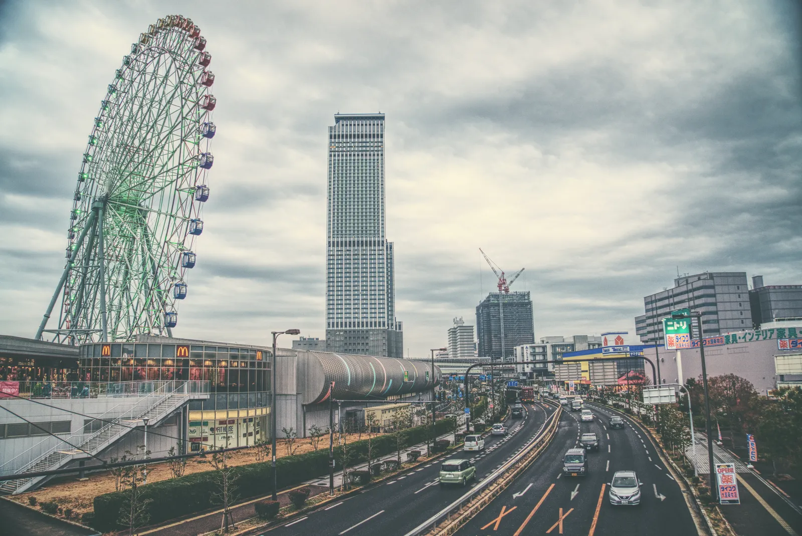 The Ferris wheel and low station buildings at Rinku Town outside Kansai airport, with a tall office tower rising behind them and a wide multi-lane road running past under a grey sky