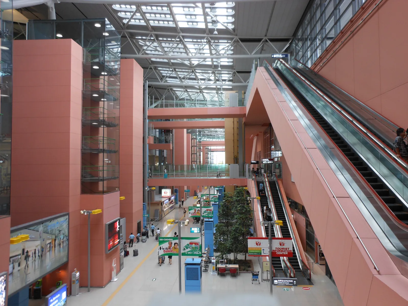Inside Terminal 1 at Kansai airport, a tall open canyon of red-panelled walls with glass lifts, long escalators and a glazed roof letting daylight down onto the check-in level far below