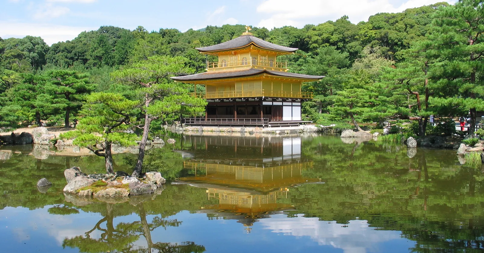 The Golden Pavilion at Kinkaku-ji in Kyoto, its upper two storeys covered in gold leaf, standing at the edge of a still pond with pines and wooded hills behind it and its reflection in the water