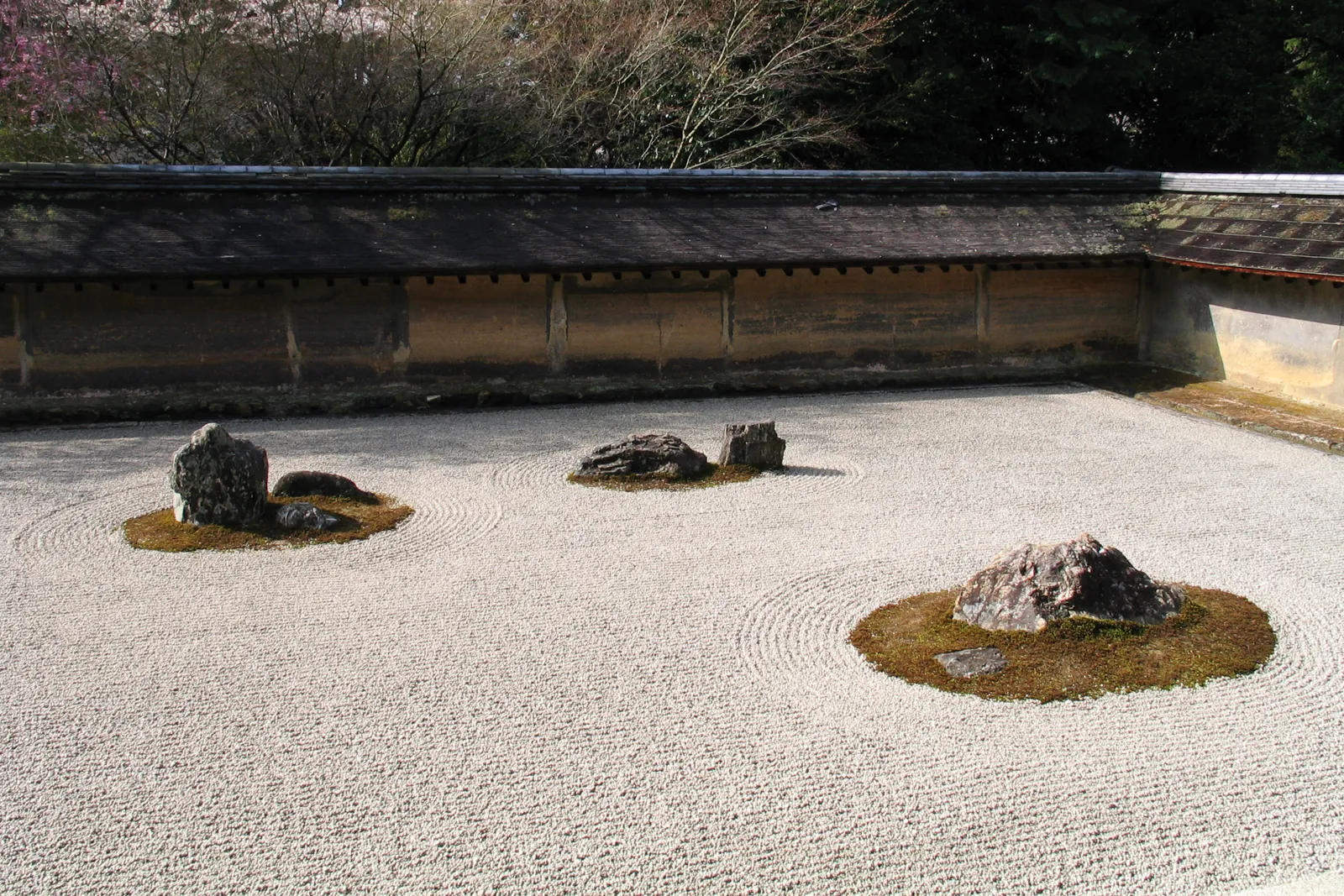 The rock garden at Ryoan-ji in Kyoto, groups of dark stones set in moss on a rectangle of raked white gravel, enclosed by a low weathered clay wall with trees above it