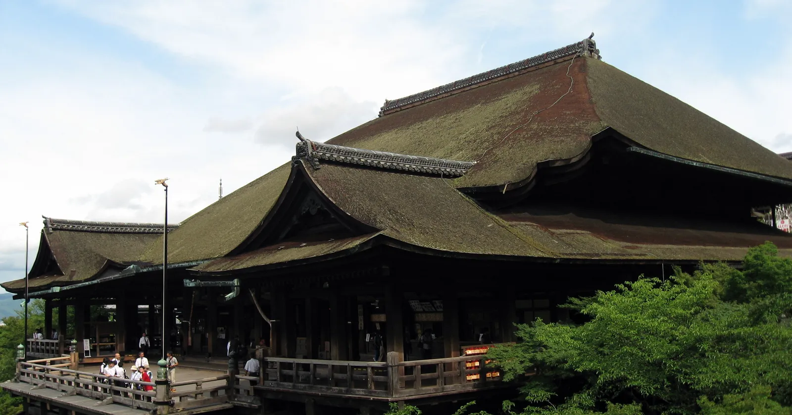 The main hall of Kiyomizu-dera in Kyoto seen from the side, a broad cypress-bark roof over a wooden veranda that runs out along the hillside with visitors standing at its rail