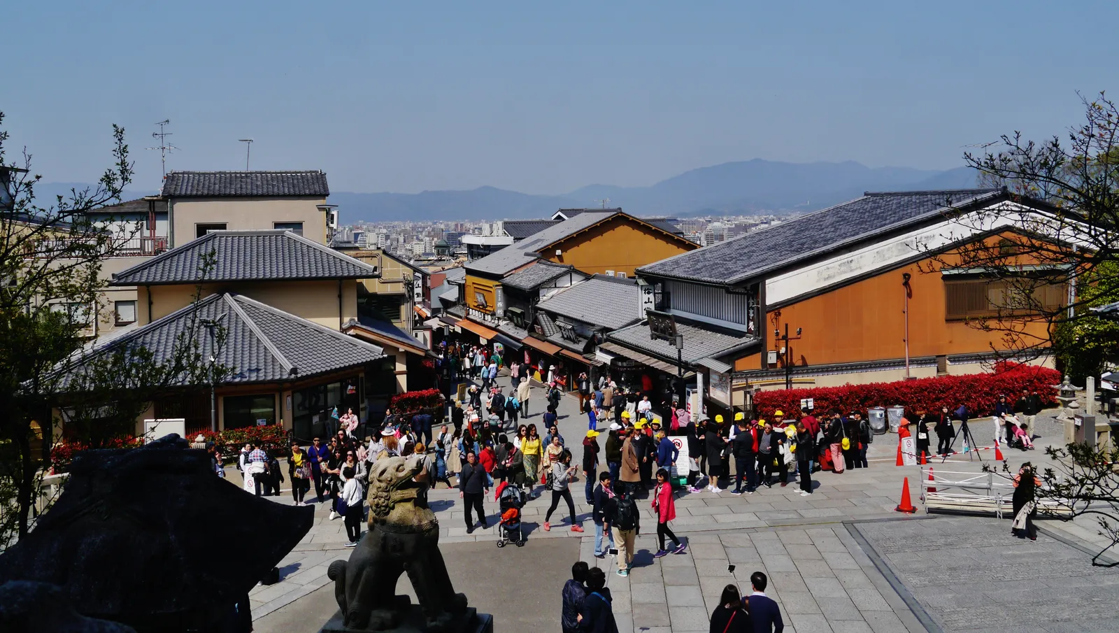 Looking down the stepped lane of Sannenzaka in Kyoto, tiled roofs and low shopfronts crowding both sides, the street full of people and the hills of the city visible in the distance