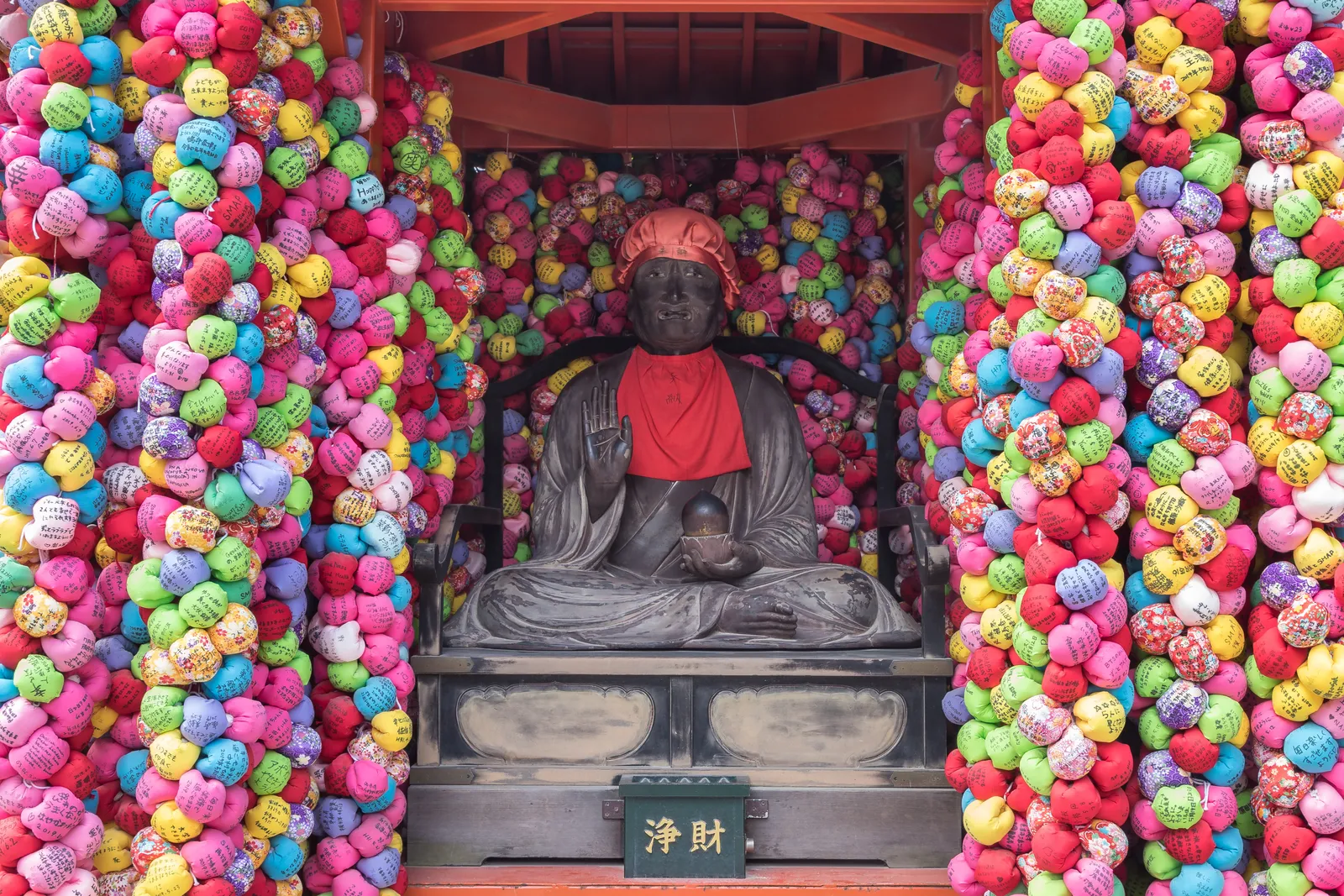 A dark seated statue at Yasaka Koshin-do in Kyoto framed on all sides by dense strings of small cloth balls in pink, blue, yellow and green hung from floor to ceiling
