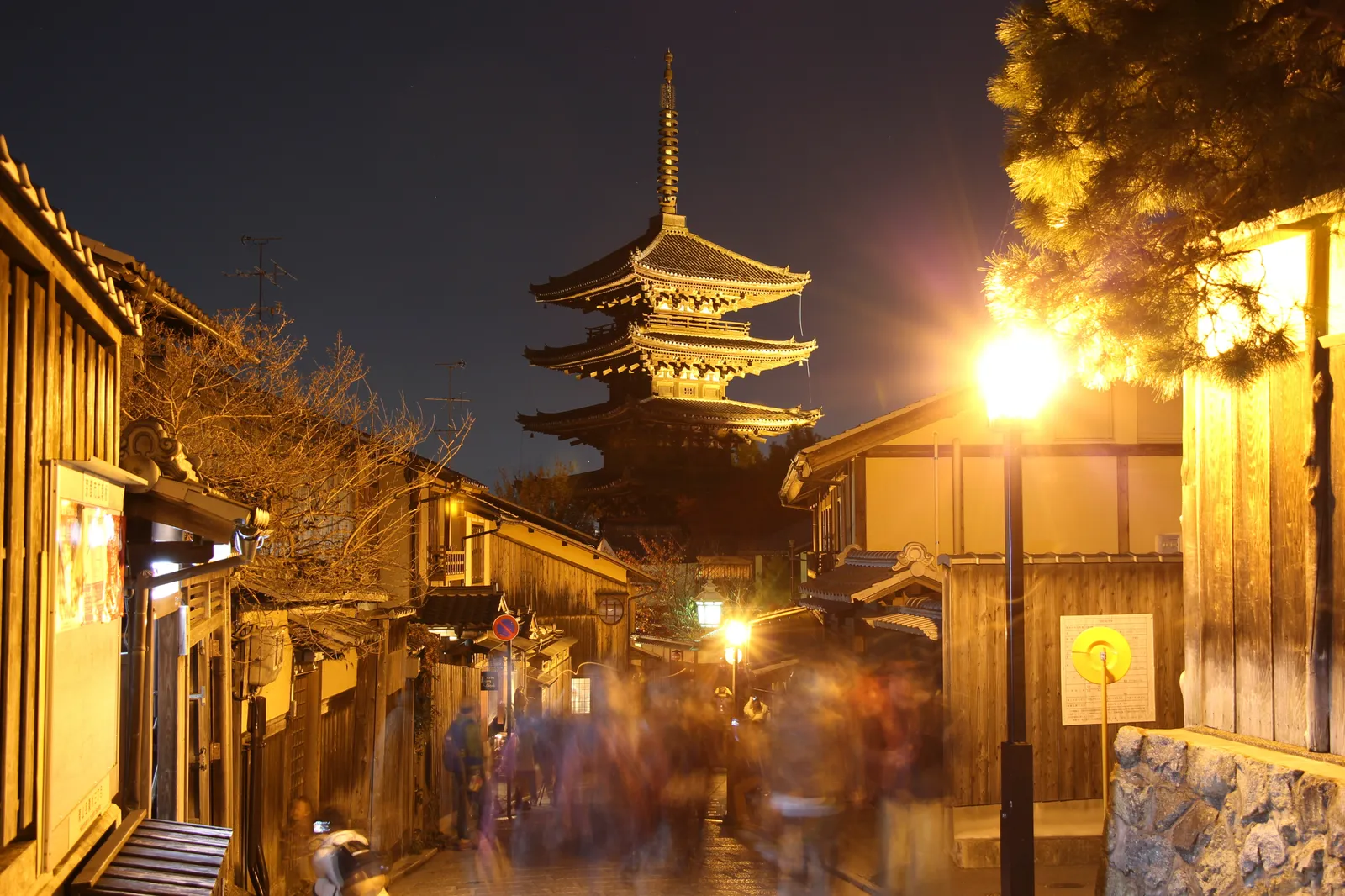 The five-storey Yasaka Pagoda lit at night above a narrow sloping lane in Kyoto, wooden shopfronts and street lanterns on both sides and people moving along the paving below it