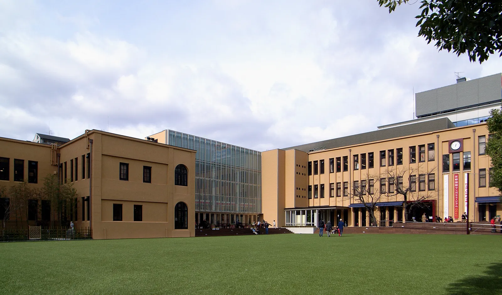The Kyoto International Manga Museum seen across its lawn, two ochre former school buildings joined by a tall glass entrance block, with people sitting on the grass in front
