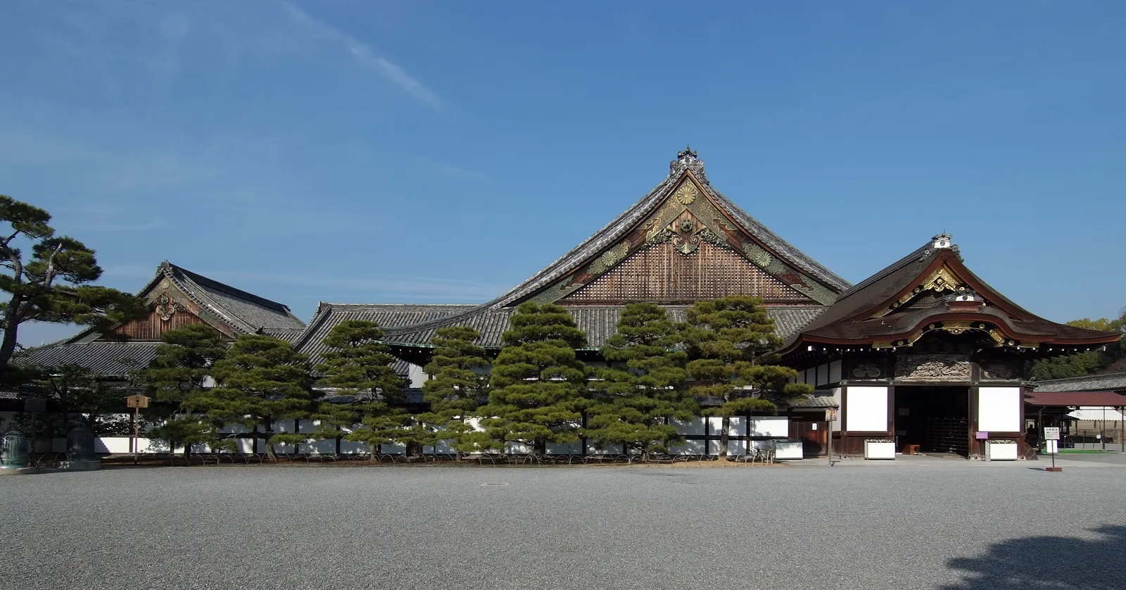 Ninomaru Palace at Nijo Castle in Kyoto seen across a wide gravel courtyard, a row of low wooden buildings with layered tiled roofs and clipped pines standing in front of them