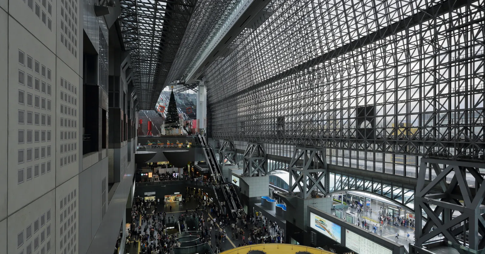 The main hall inside Kyoto Station, a vast concourse under an exposed steel lattice roof with a glass wall rising the full height of the building and escalators climbing the levels along one side