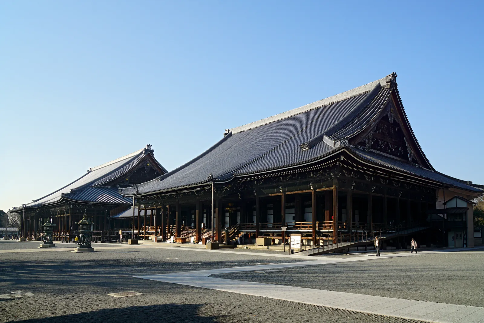 The two main halls at Nishi Honganji in Kyoto standing side by side across a wide gravel courtyard under a clear sky, each with a broad sweeping tiled roof carried on wooden pillars