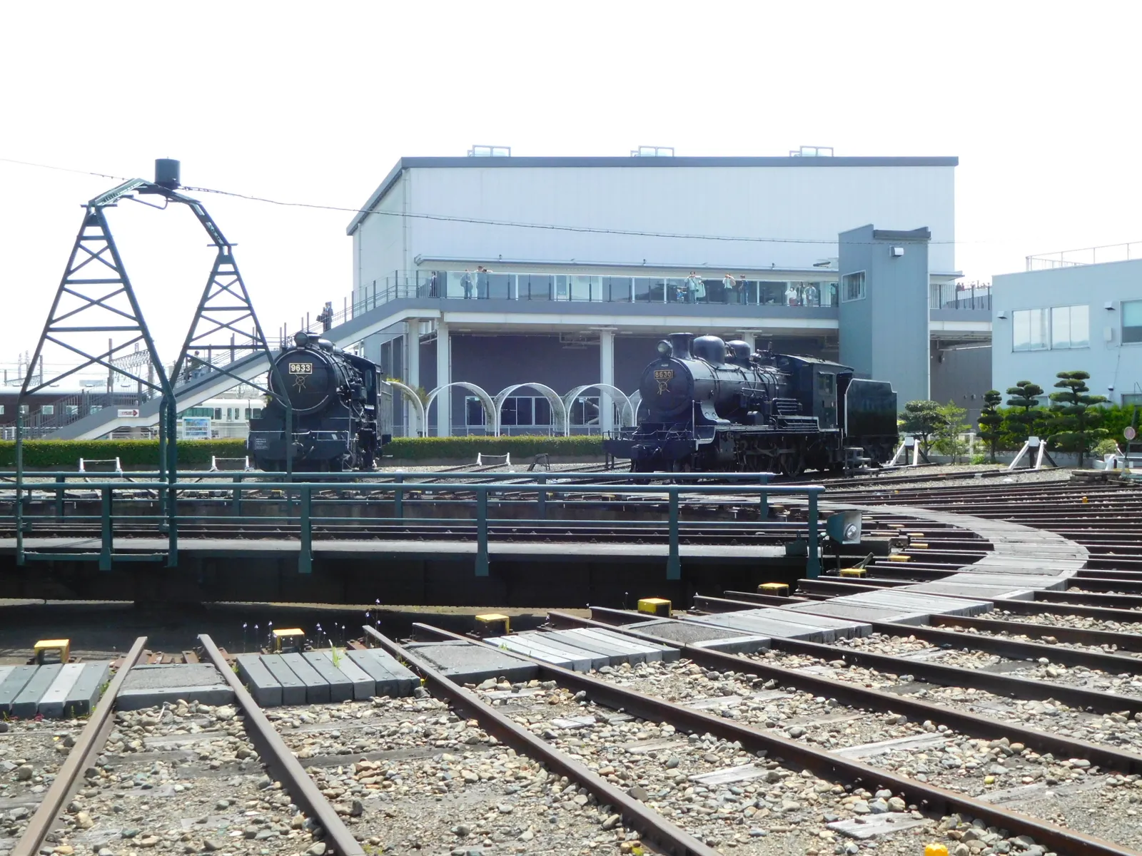 The roundhouse at the Kyoto Railway Museum, black steam locomotives standing on tracks that fan out from a turntable in the foreground towards an arched engine shed behind