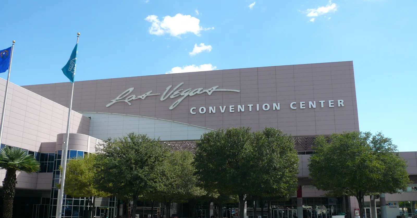 The Las Vegas Convention Center's long grey frontage with its script logo and CONVENTION CENTER lettering across the top, flags and trees along the approach under a blue sky