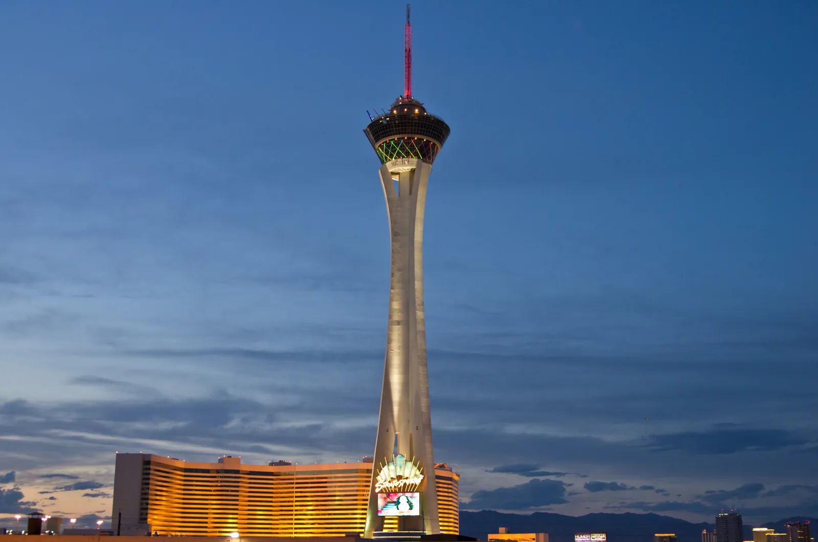 The Strat's slender concrete observation tower at dusk in Las Vegas, its pod lit at the top above the low curved casino building at its base