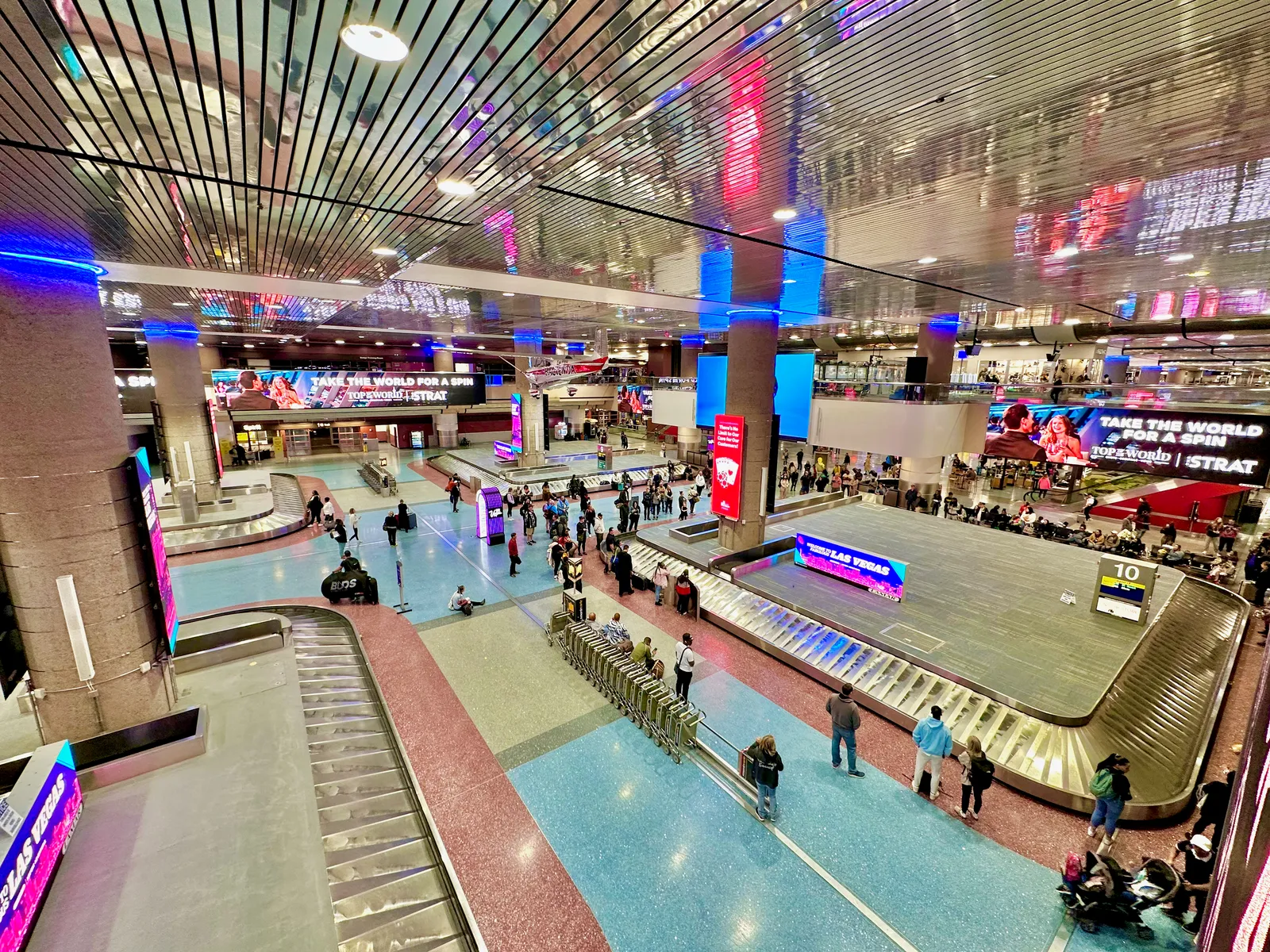 A baggage claim hall at Harry Reid International Airport seen from the level above, its carousels ringed by waiting passengers under a ceiling hung with neon tubes and video screens