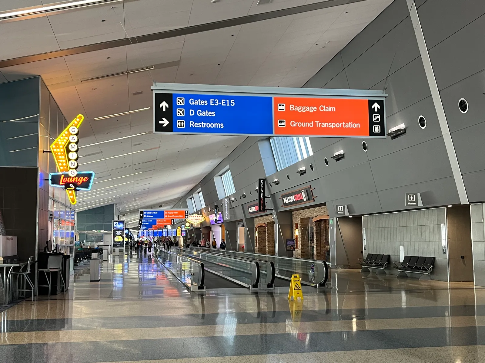 A wide concourse at Harry Reid International Airport with a moving walkway down the middle and an overhead sign pointing to gates E3 to E15, the D gates, baggage claim and ground transportation