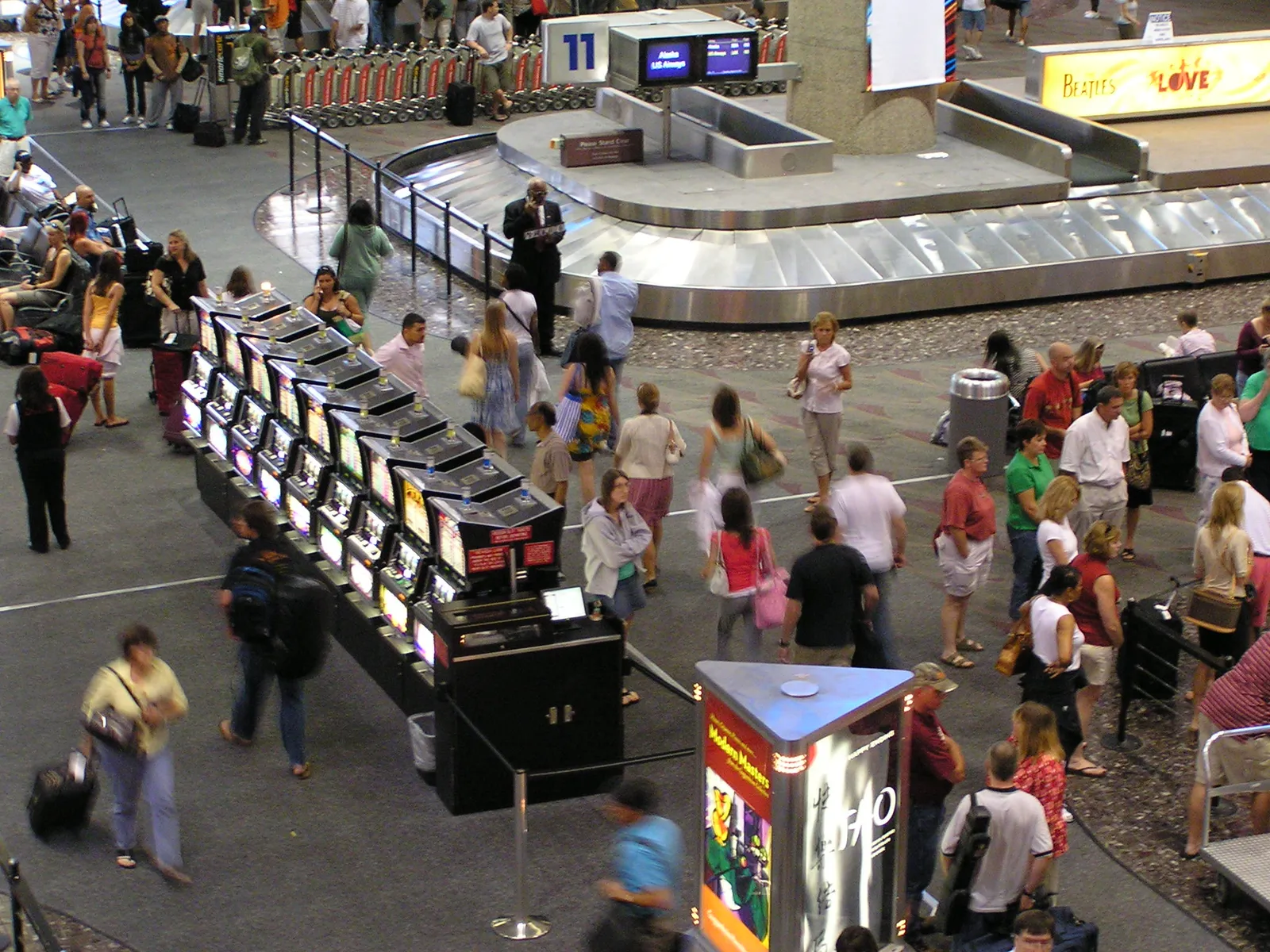 A long bank of slot machines standing beside a baggage carousel at the Las Vegas airport, with arriving passengers walking past them