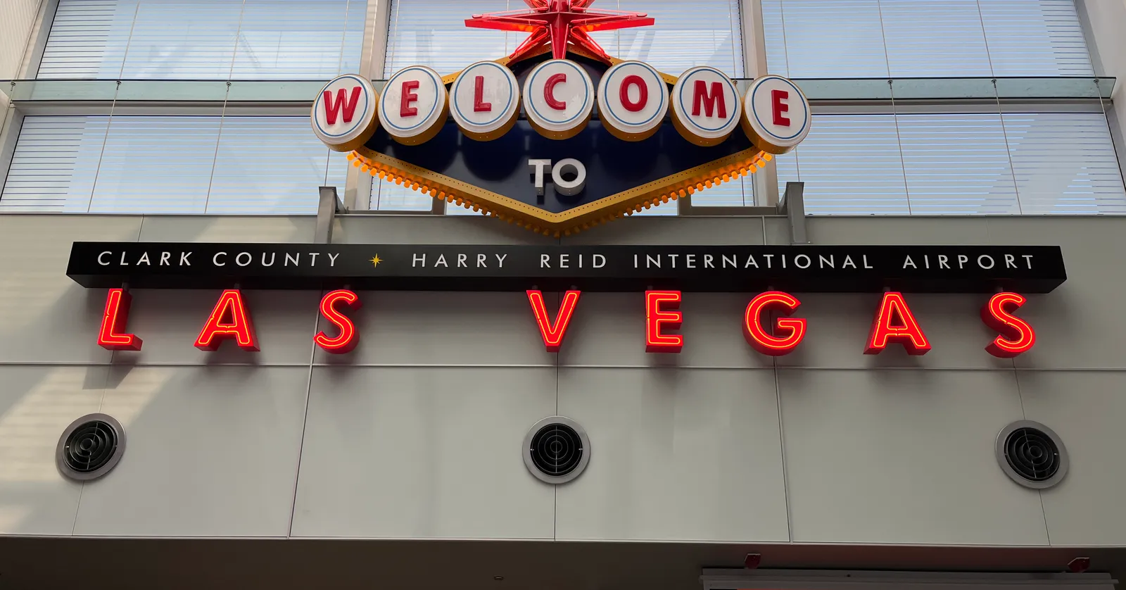 A neon Welcome to Las Vegas sign mounted high inside Harry Reid International Airport, reading Clark County Harry Reid International Airport above red LAS VEGAS lettering