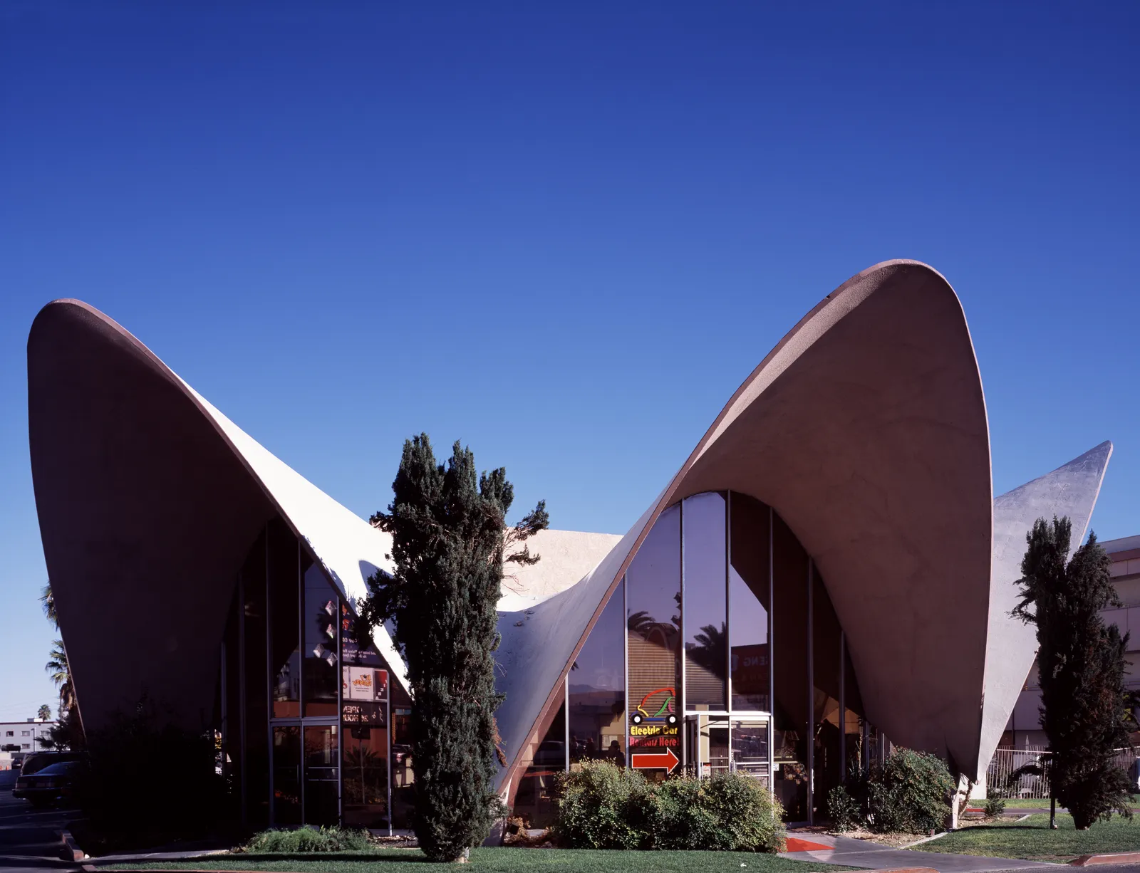 The La Concha Motel lobby, a white concrete shell of three curved sweeping points over a glass frontage, against a deep blue sky with cypress trees in front