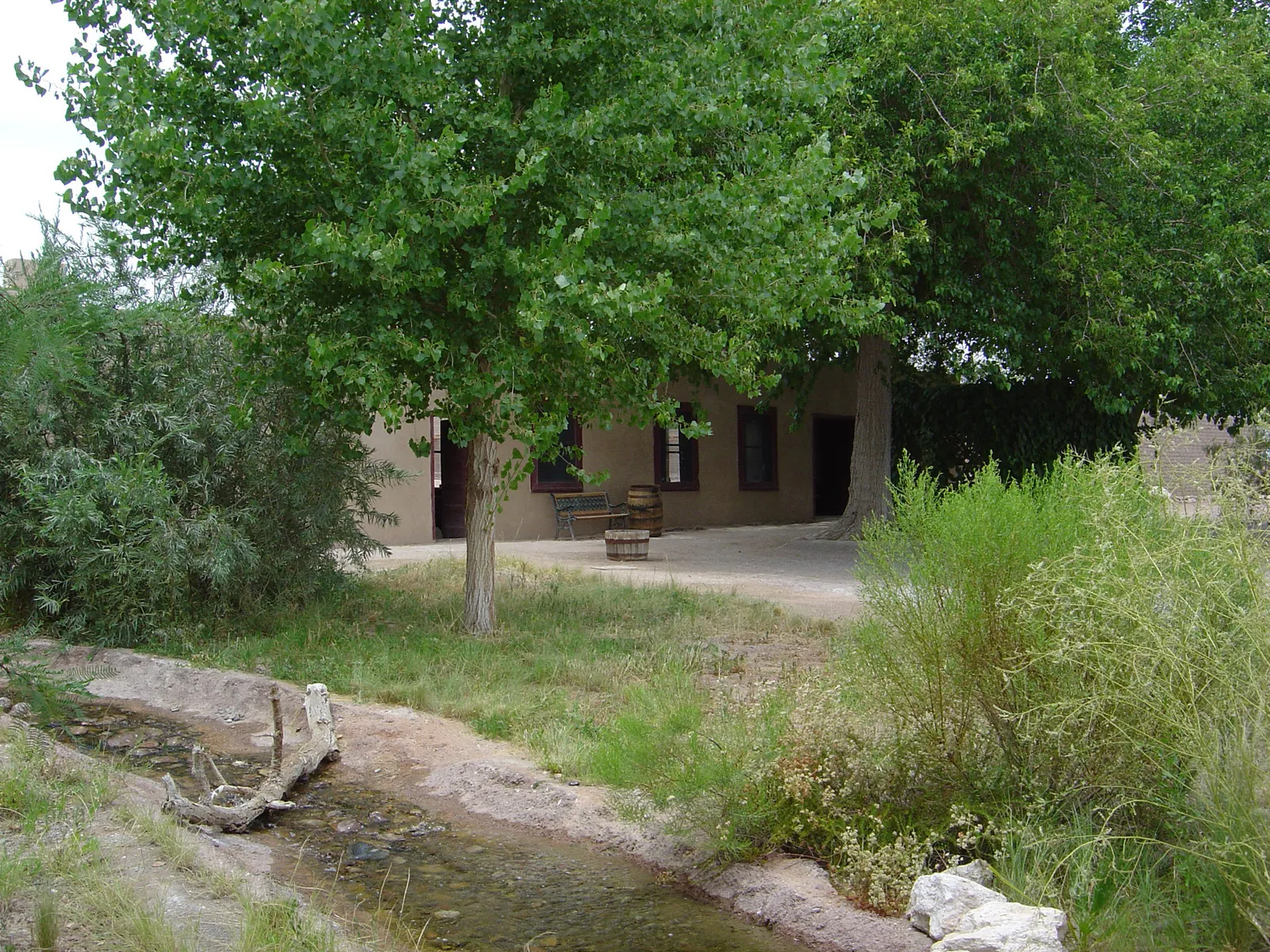 The surviving adobe building of the Old Las Vegas Mormon Fort under mature cottonwood trees, with a water channel running through the grass in front of it