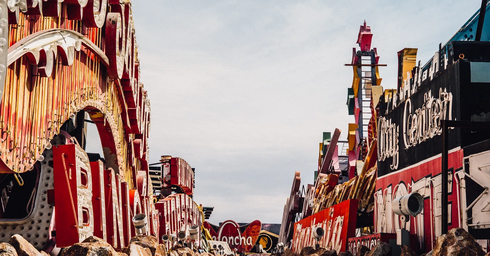 A gravel lane running between two banked rows of retired Las Vegas neon signs standing upright in the Neon Museum's boneyard under a pale sky