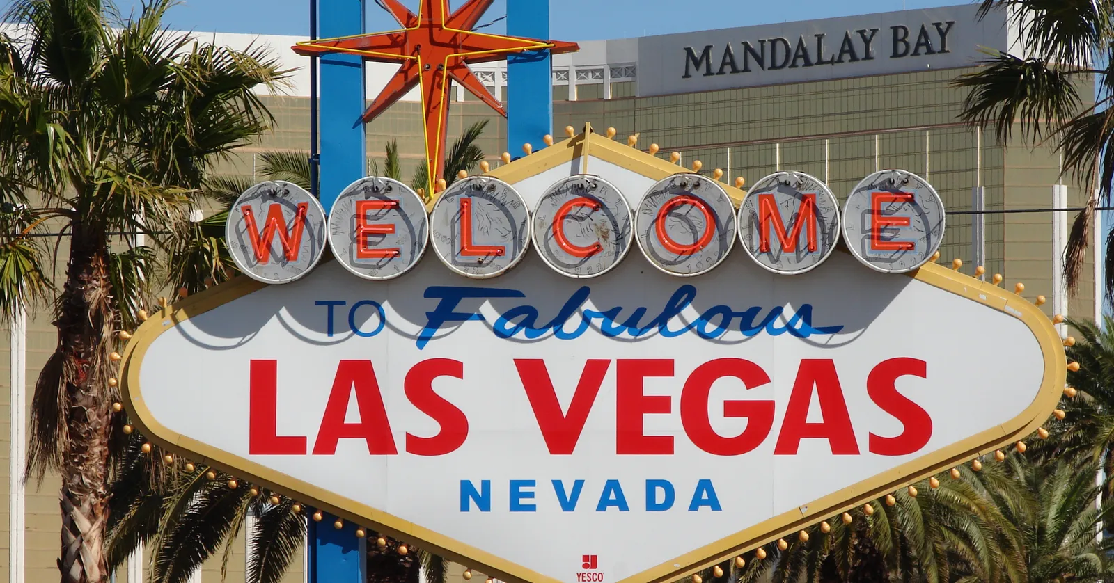 The Welcome to Fabulous Las Vegas Nevada sign, a white diamond with red and blue lettering and a red star on top, framed by palm trees with the Mandalay Bay resort behind it