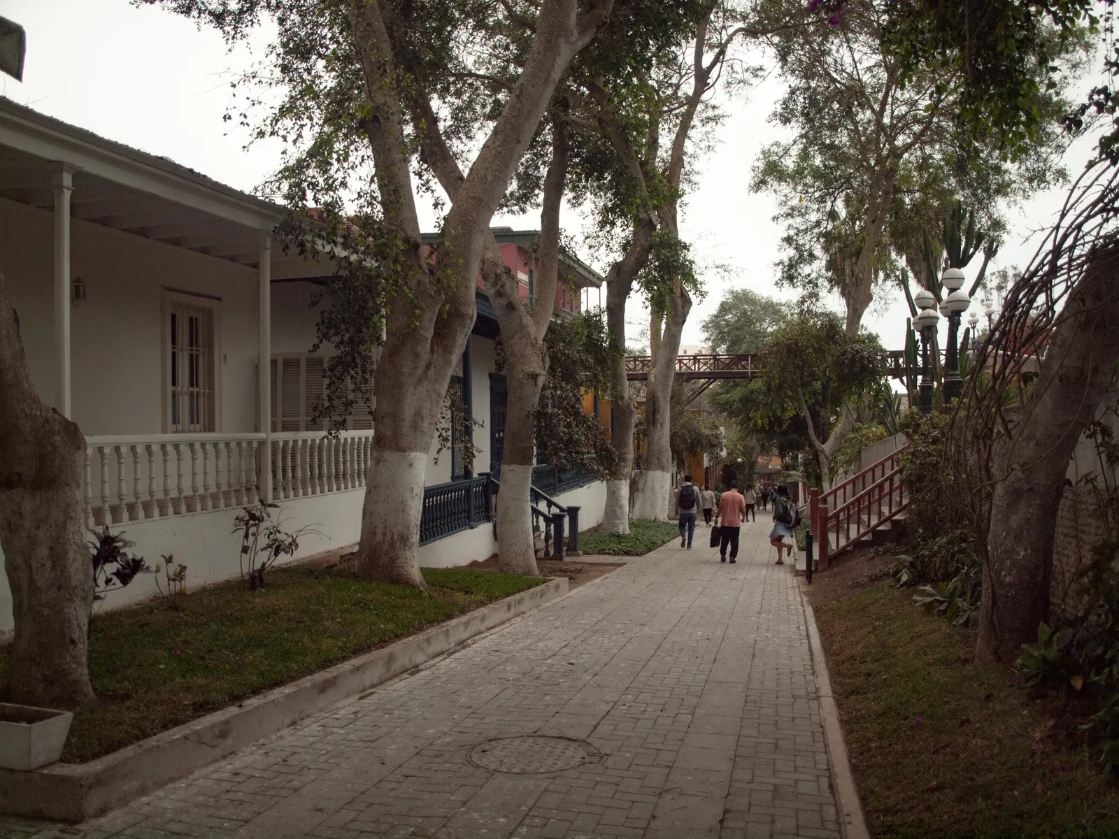 The paved Bajada de Baños in Barranco, Lima, running downhill between old white timber houses and mature trees with the wooden footbridge crossing overhead