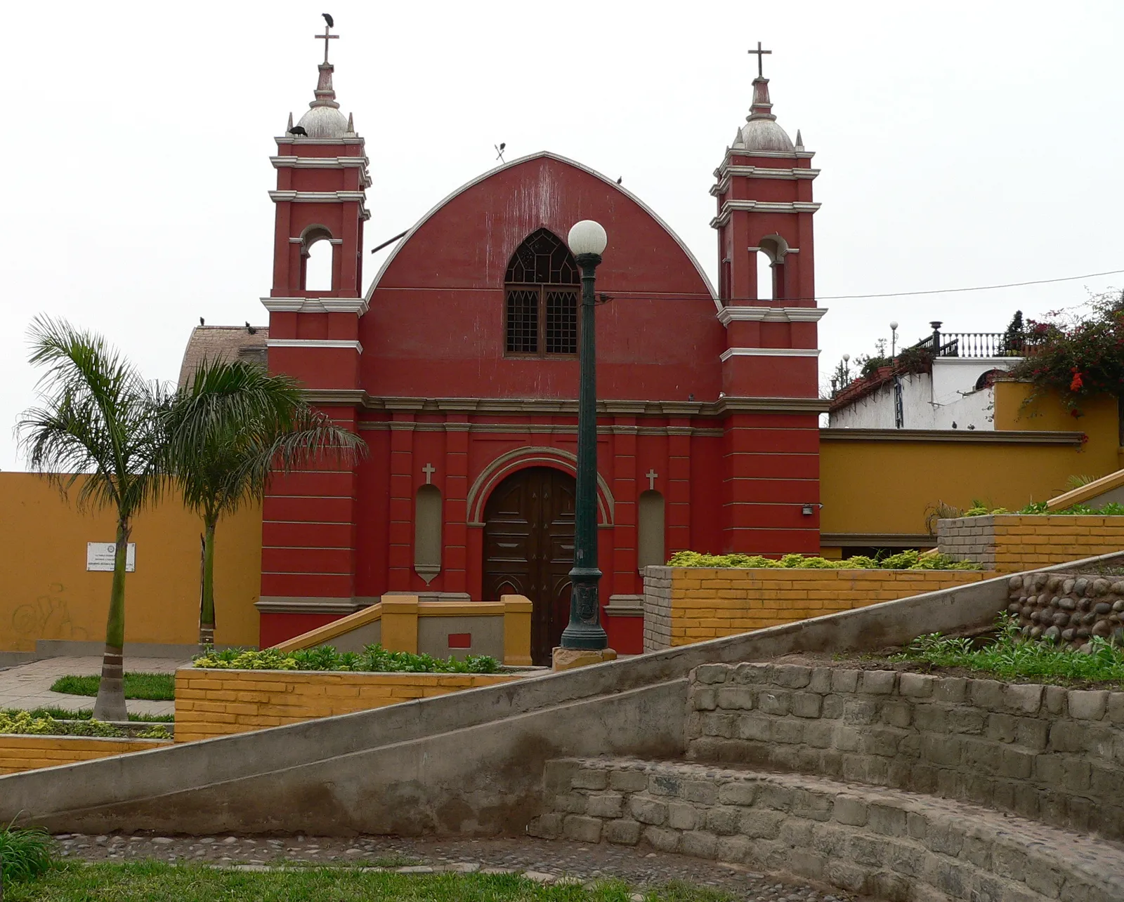 The red adobe front of La Ermita in Barranco, Lima, with two small bell towers, a pointed gable and a boarded wooden door, seen across low garden terraces