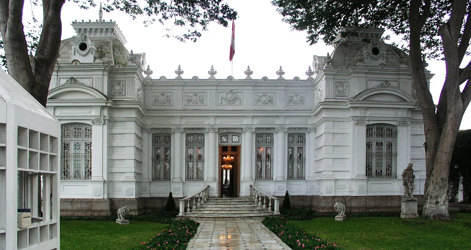 The white French-styled front of the Museo Pedro de Osma in Barranco, Lima, with a Peruvian flag over the pediment, stone lions on the steps and a flower bed running up to the door